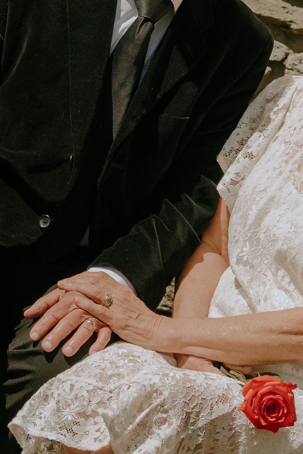 Elderly couple holding hands at wedding, bride in lace dress with red rose, groom in dark suit with ring visible.