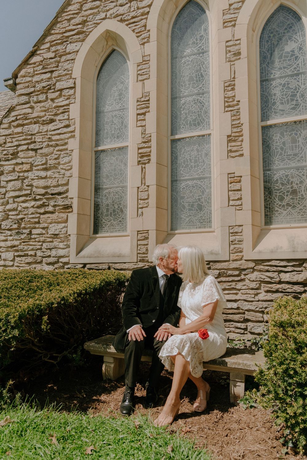 Elderly couple sharing a romantic kiss on a stone bench outside a historic stone church with arched stained glass windows.
