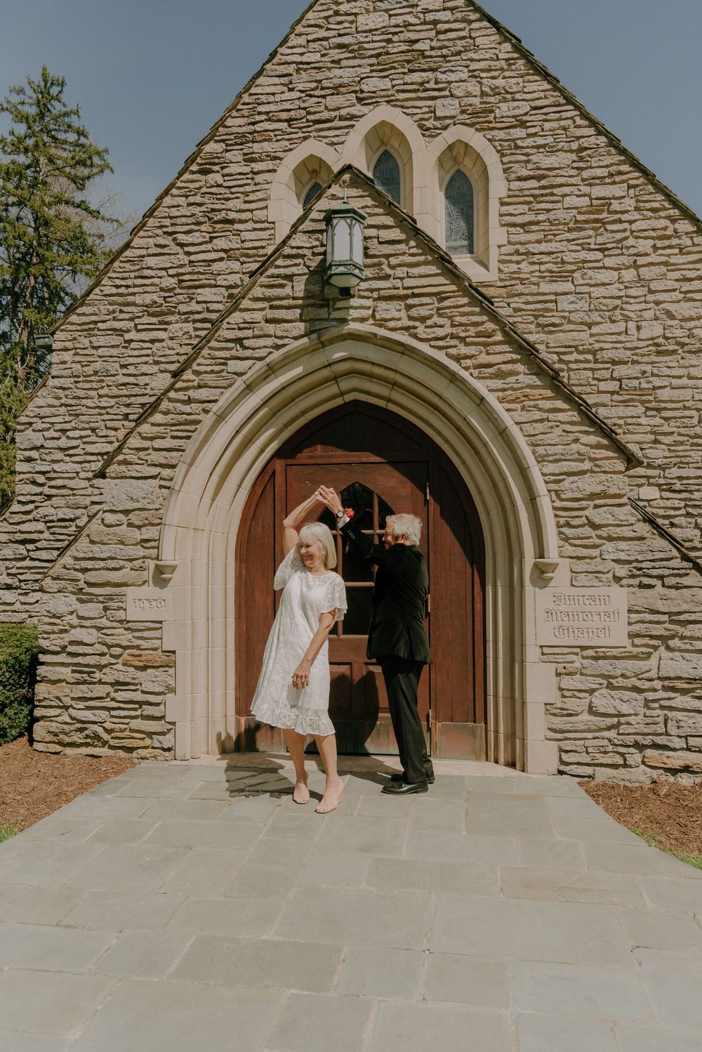 Elderly couple dancing outside a stone church with Gothic arch doorway on their wedding day.