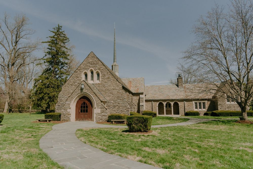 Stone church with Gothic arched entrance, steeple, and lush green lawn surrounded by bare trees on a cloudy day.