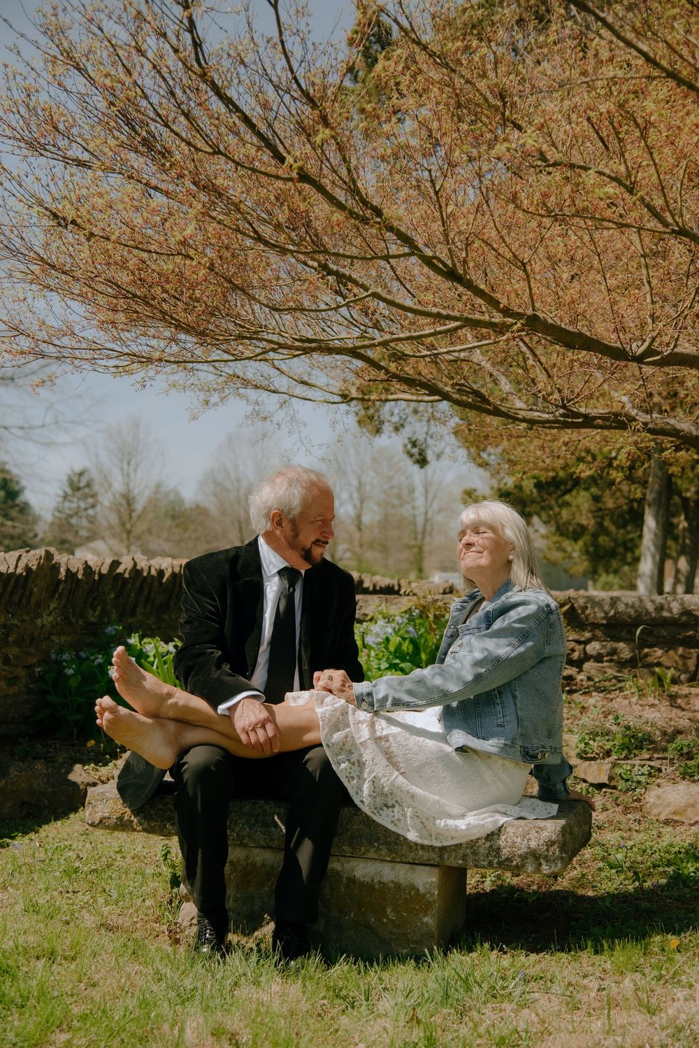 An elderly couple sits on a stone bench under a tree, smiling at each other on a sunny day.