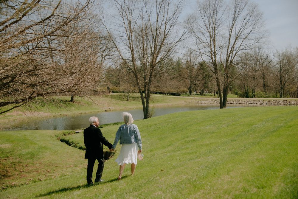 Elderly couple holding hands walking on lush green grass near a pond surrounded by bare trees in spring.