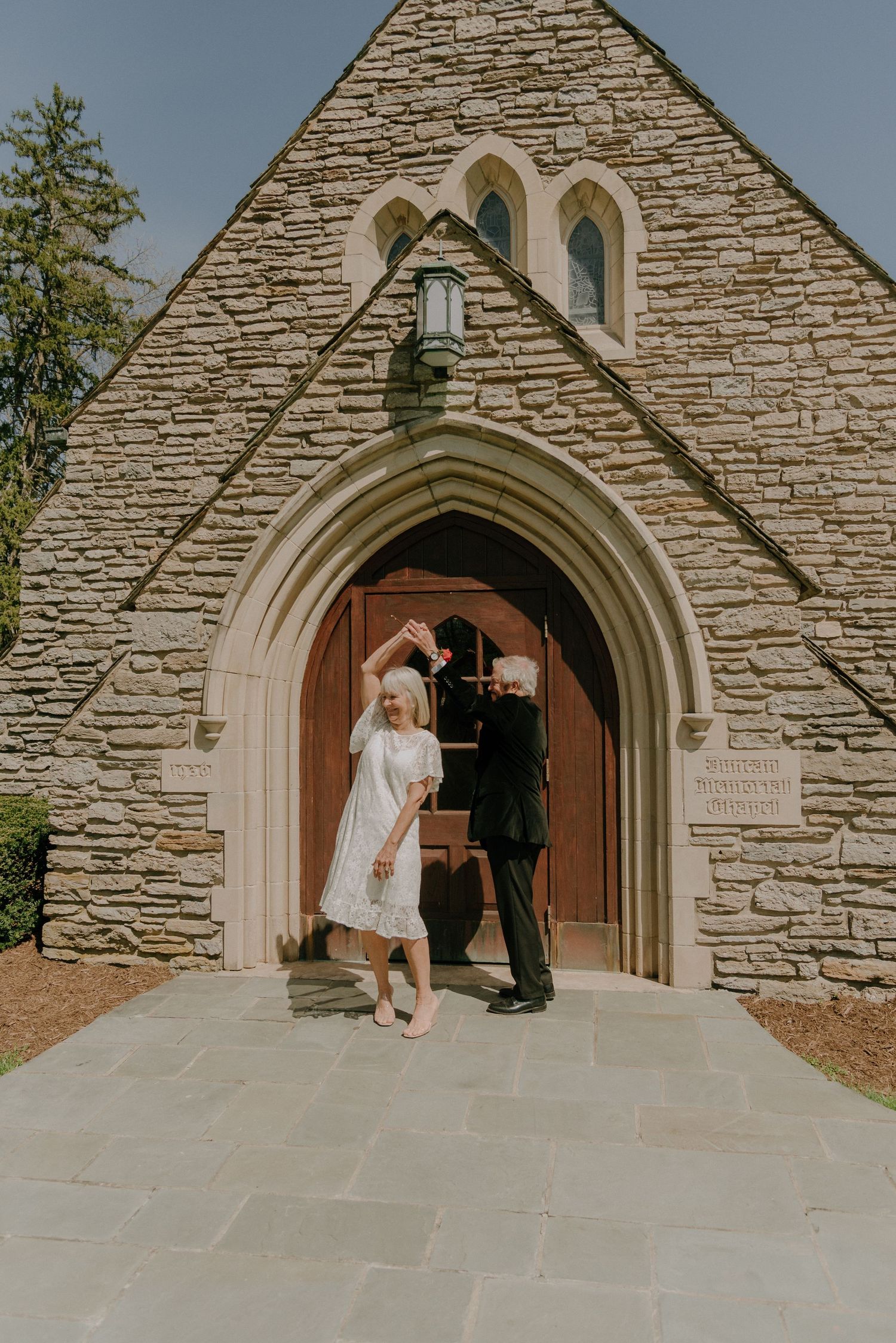 Elderly couple dancing outside a stone church in wedding attire, bride in white dress, groom in black suit.