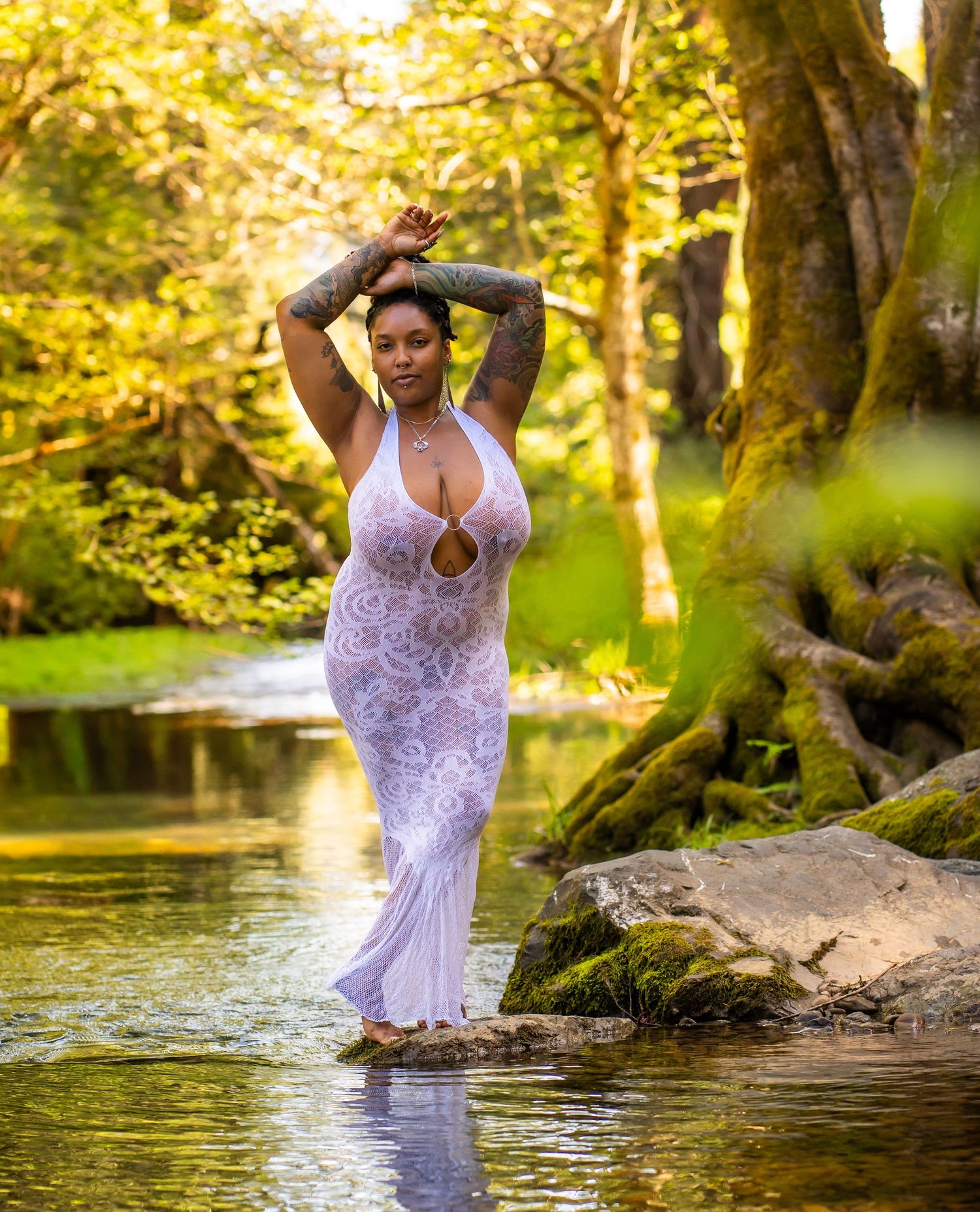 Woman in sheer white lace dress posing gracefully in a serene forest stream surrounded by lush greenery.