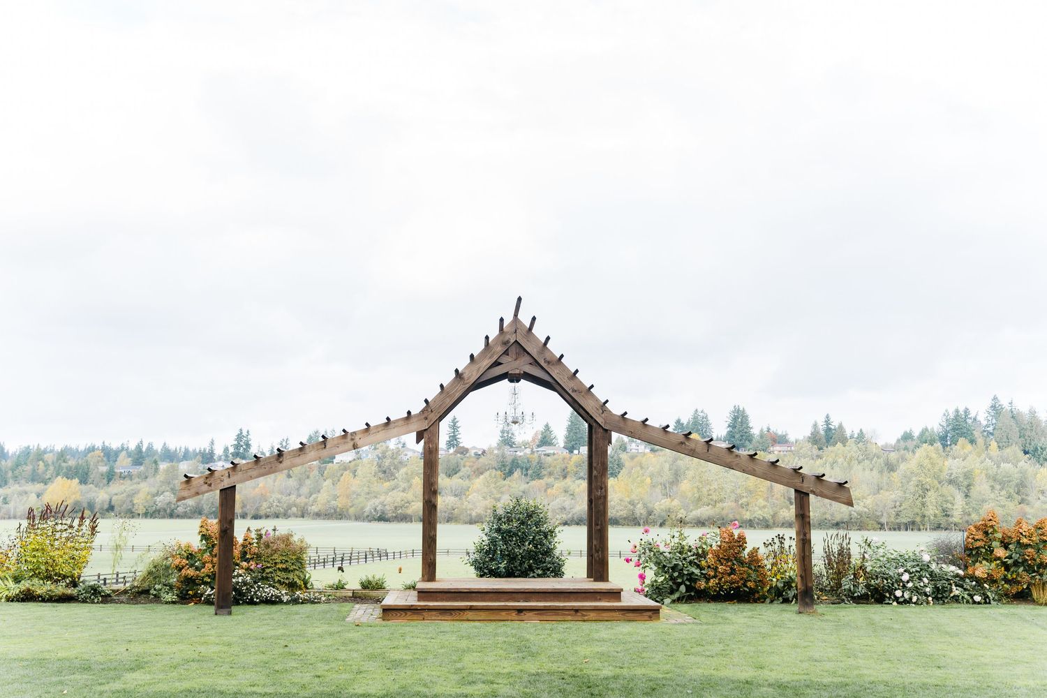 Rustic wooden wedding arch on lush green lawn with garden backdrop and misty trees at outdoor venue.