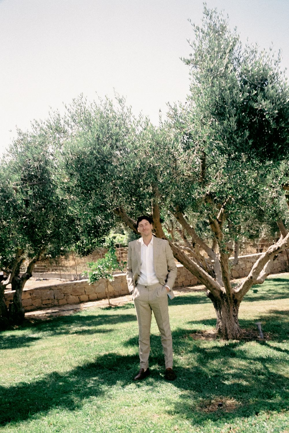 Groom in beige suit standing confidently under olive trees on green lawn at outdoor wedding venue.