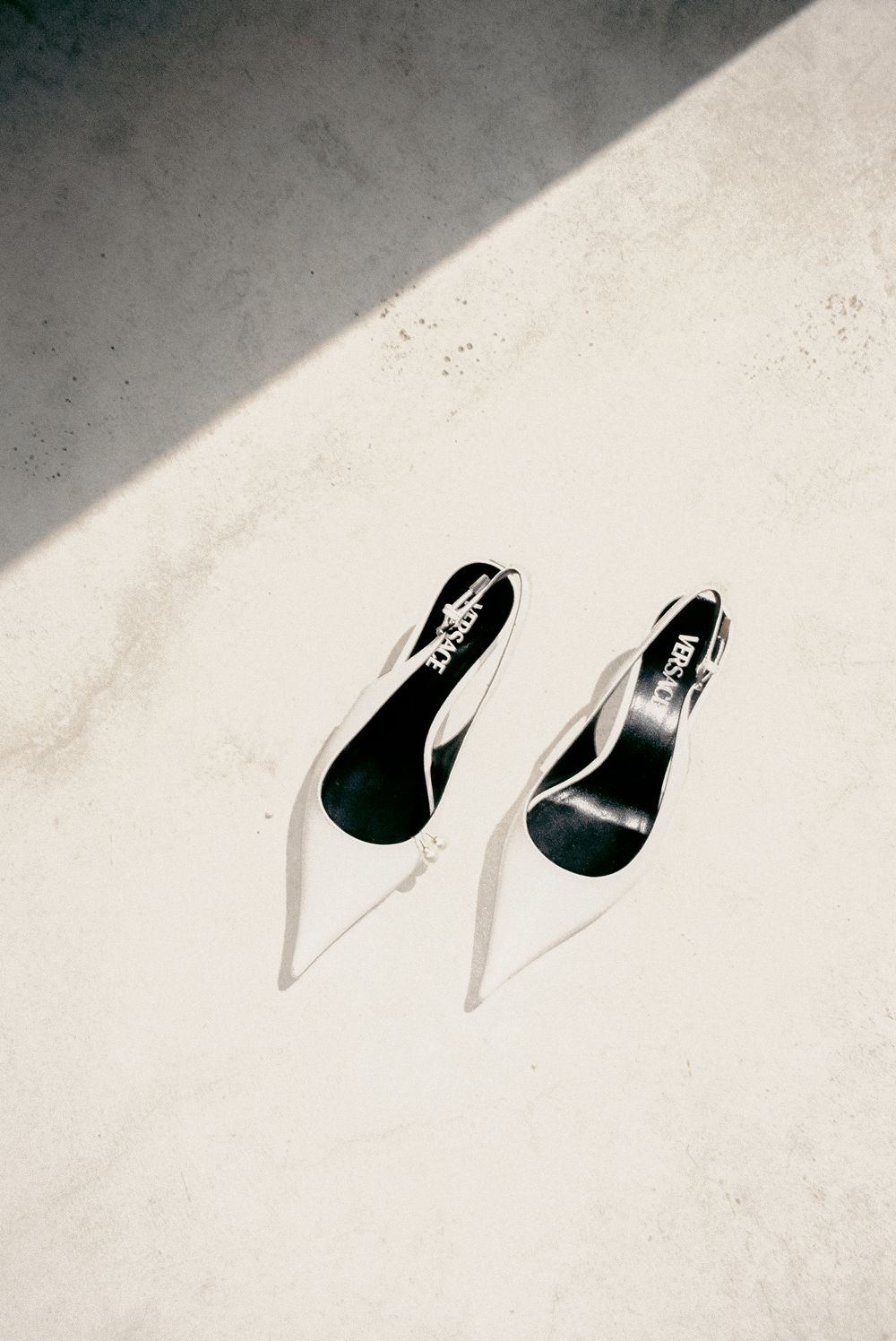 White slingback heels with black insoles casting shadows on a bright concrete surface in natural sunlight.