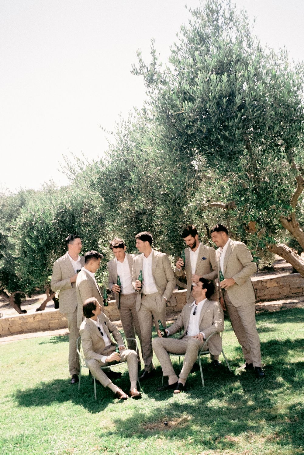 Groomsmen in tan suits gather under olive trees on a sunny day, some seated, holding drinks at an outdoor wedding.