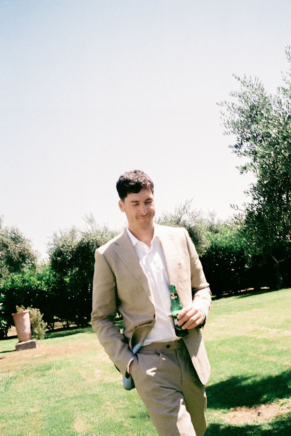 Handsome groom in beige linen suit holding a green bottle while standing on grass surrounded by olive trees.