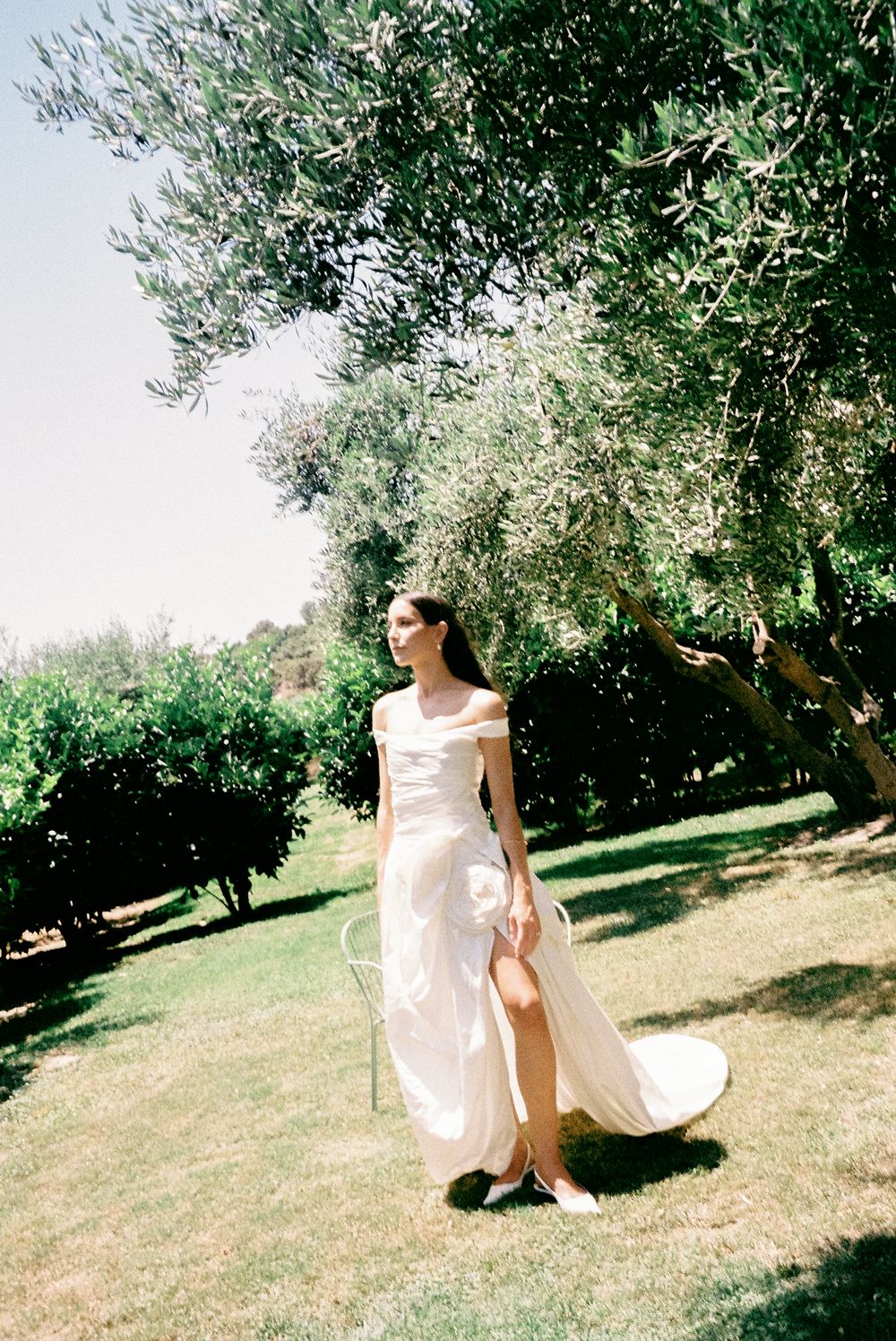 Bride in elegant off-shoulder white wedding gown with rose detail walks through lush garden.