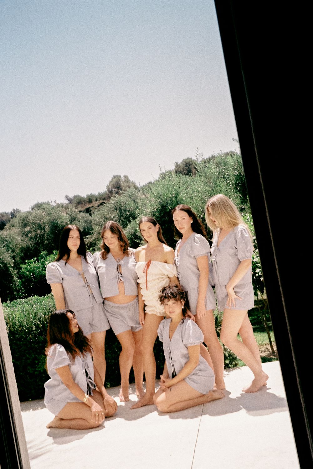 Seven bridesmaids in matching gray pajama sets pose together outdoors in sunny garden setting.