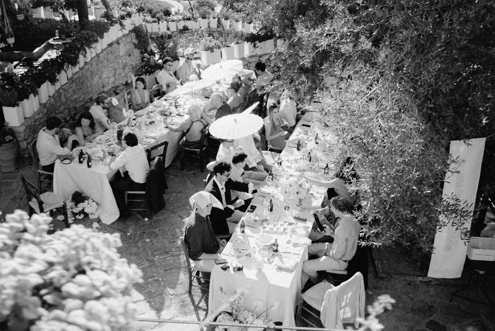 Black and white aerial view of wedding guests dining at long decorated tables in a lush garden setting.