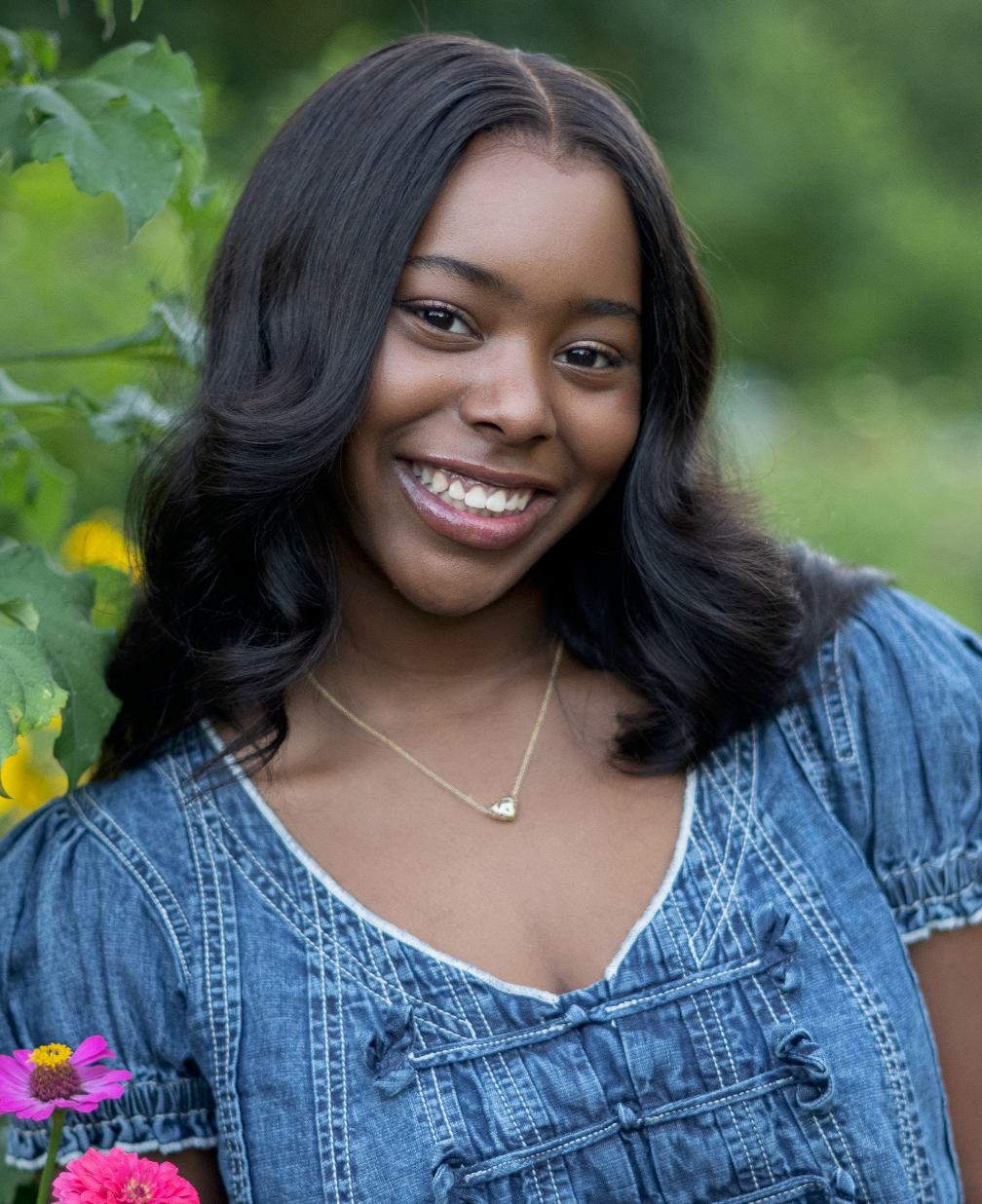 Smiling young woman in denim top wearing a gold necklace, posing outdoors with colorful flowers and green foliage.
