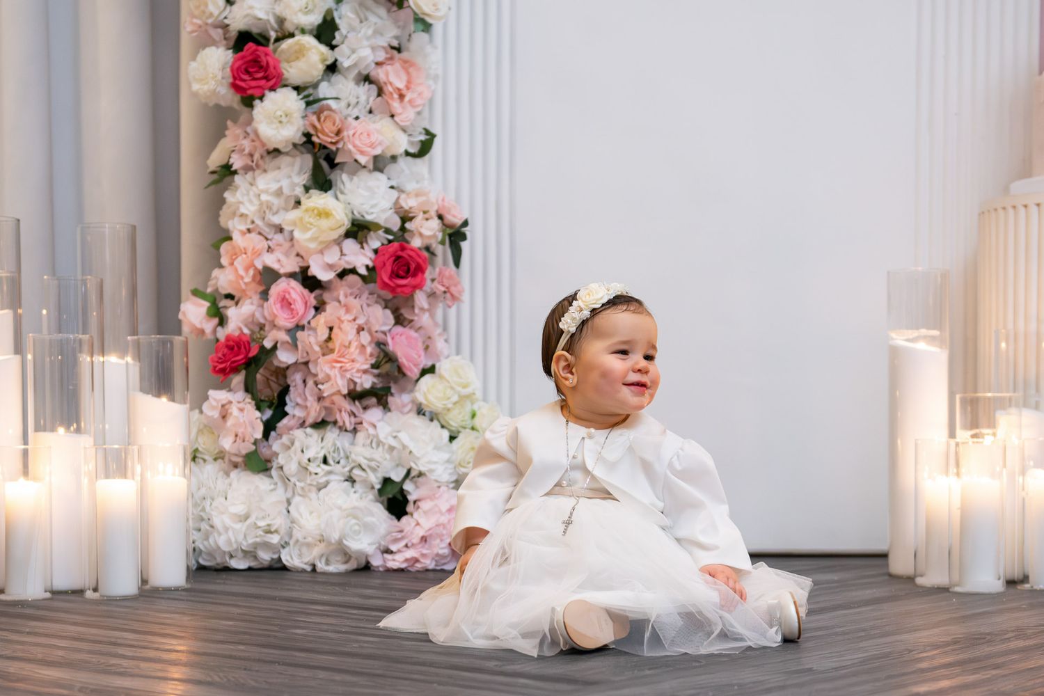 A baby girl in a white christening dress sits surrounded by candles and floral arrangements in a studio setting.