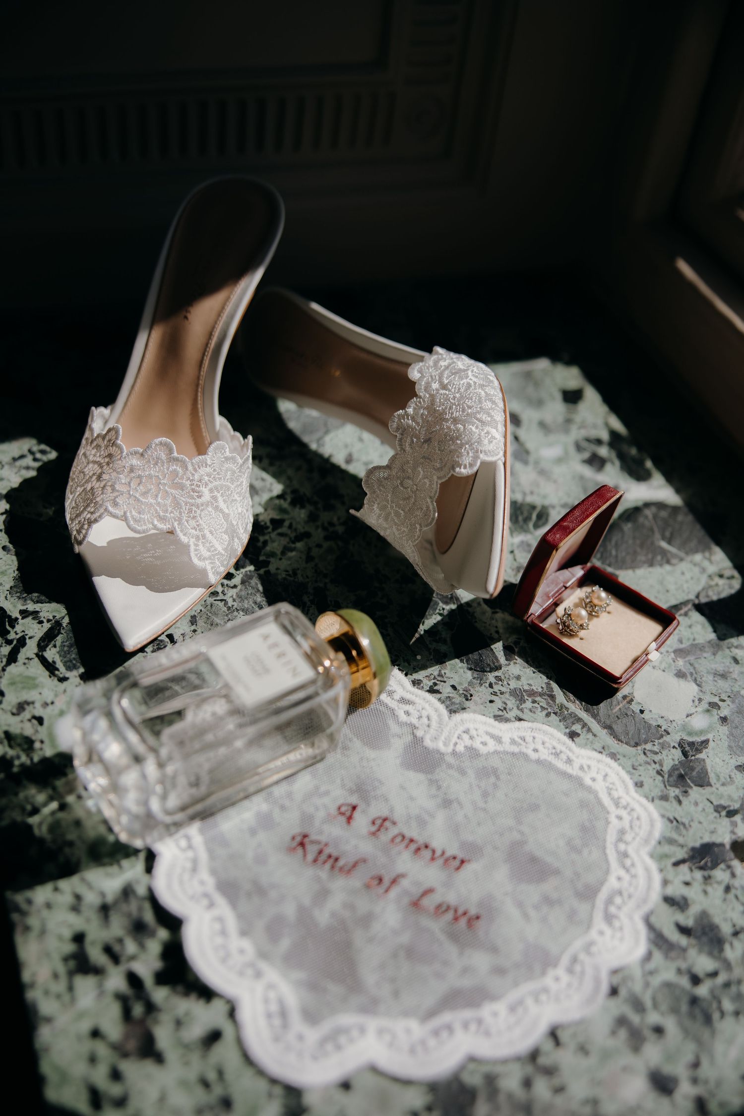 White lace bridal heels, perfume bottle, ring box, and embroidered handkerchief on marble surface for wedding day details.