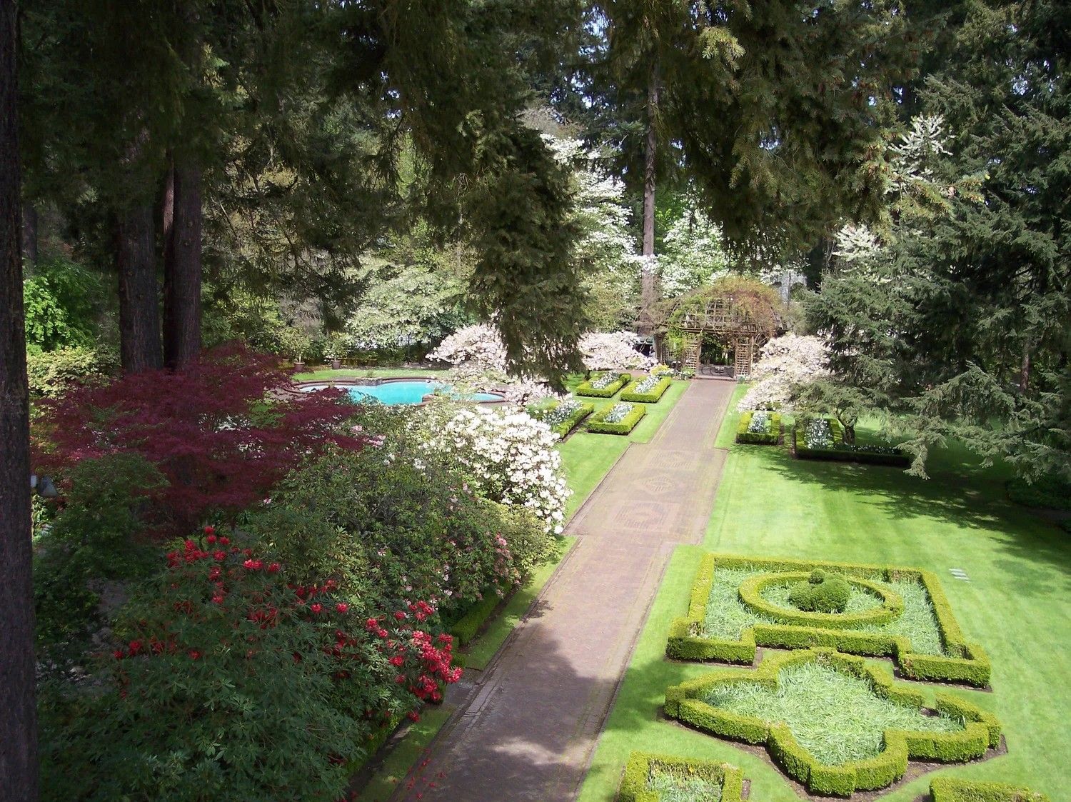 Formal English garden with manicured hedges, red maples, white flowering shrubs, and a brick pathway leading to a fountain.