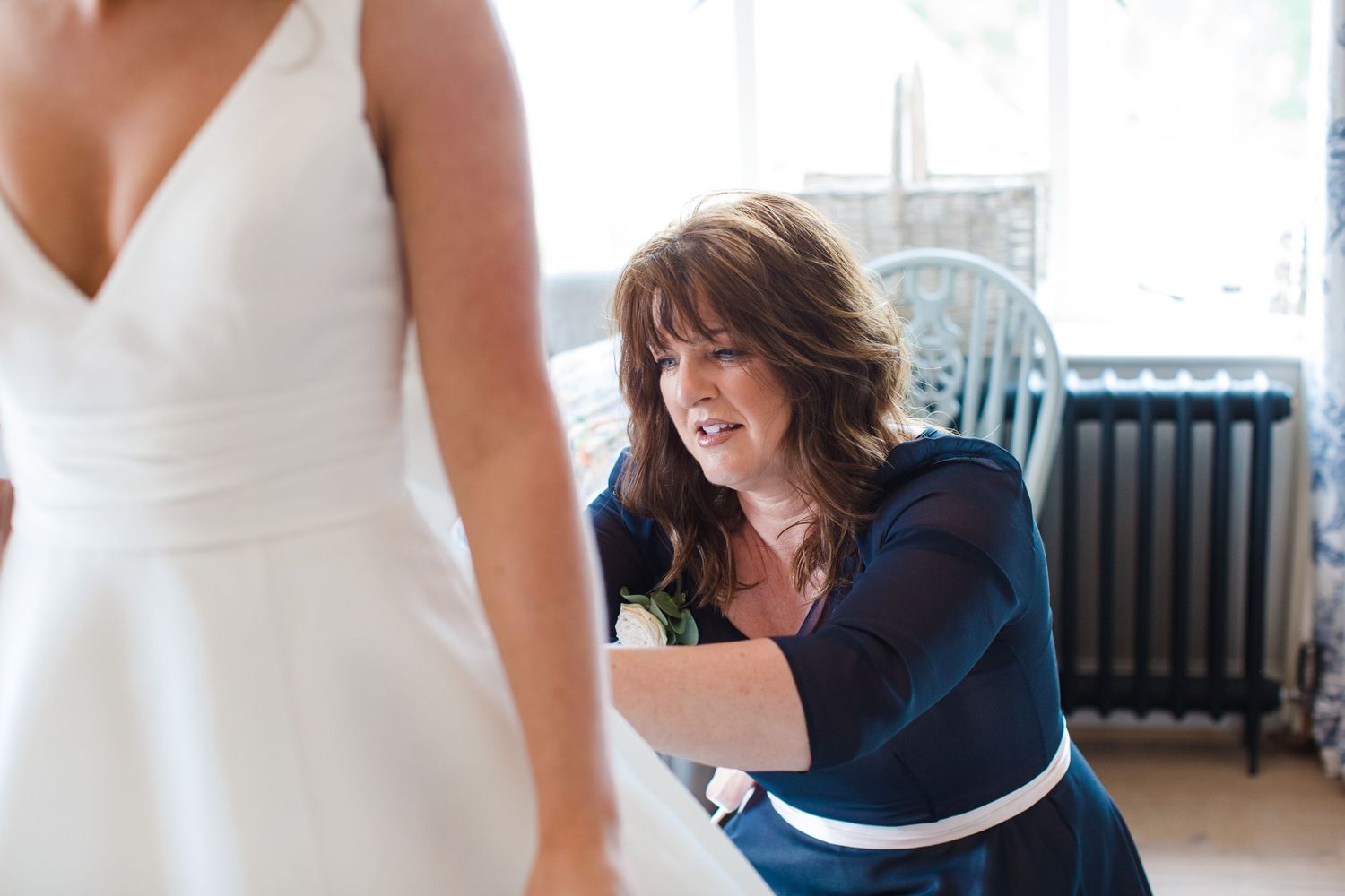 A woman in a blue dress carefully adjusts the back of a bride's white wedding gown indoors.