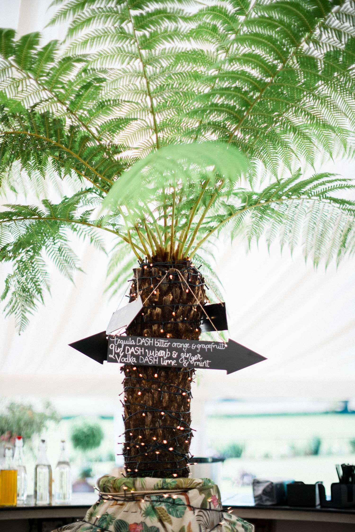 Decorative fern tree centerpiece with fairy lights and chalkboard arrow signs at an elegant tented wedding reception Oceanstone Resort