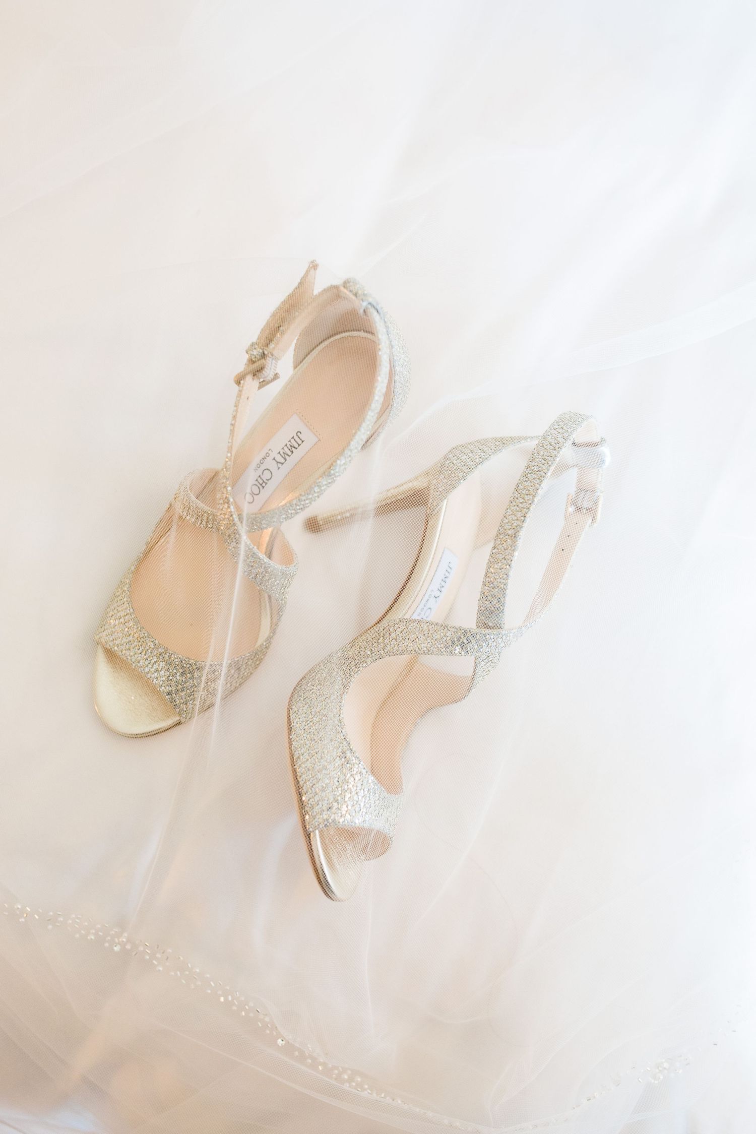 Elegant Jimmy Choo gold glitter strappy heeled sandals resting on white bridal veil fabric at Bull Point Estate