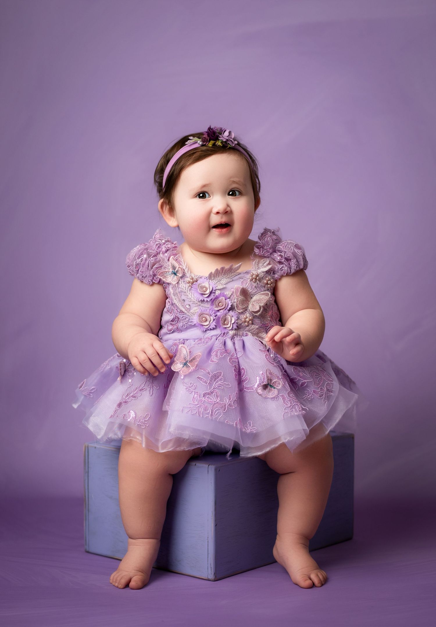 Two baby girls in yellow and purple rompers sit together in a colorful flower garden with a white picket fence.