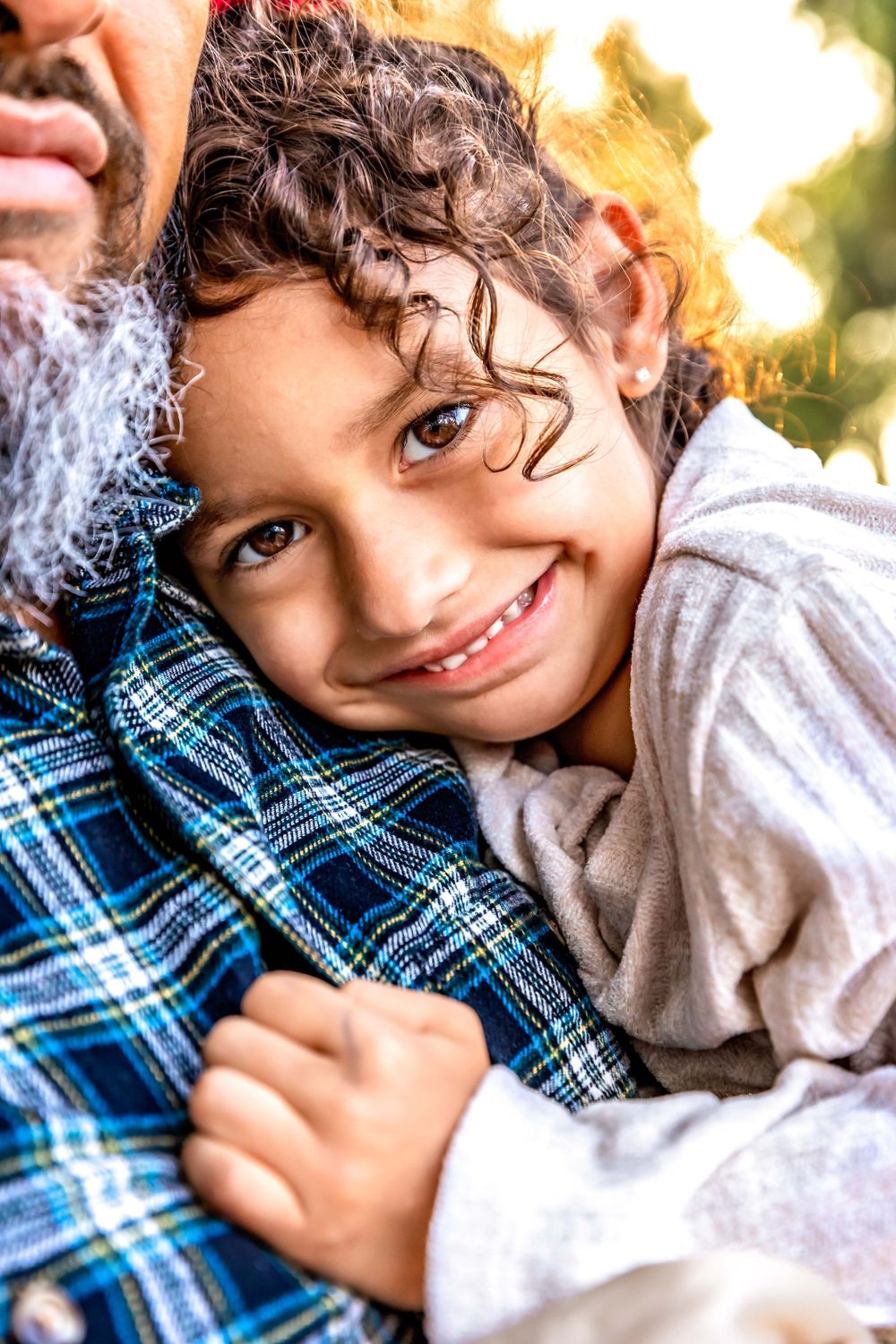A young child with curly hair rests against someone wearing a blue plaid shirt while smiling warmly taken by local photographer in Fresno,ca