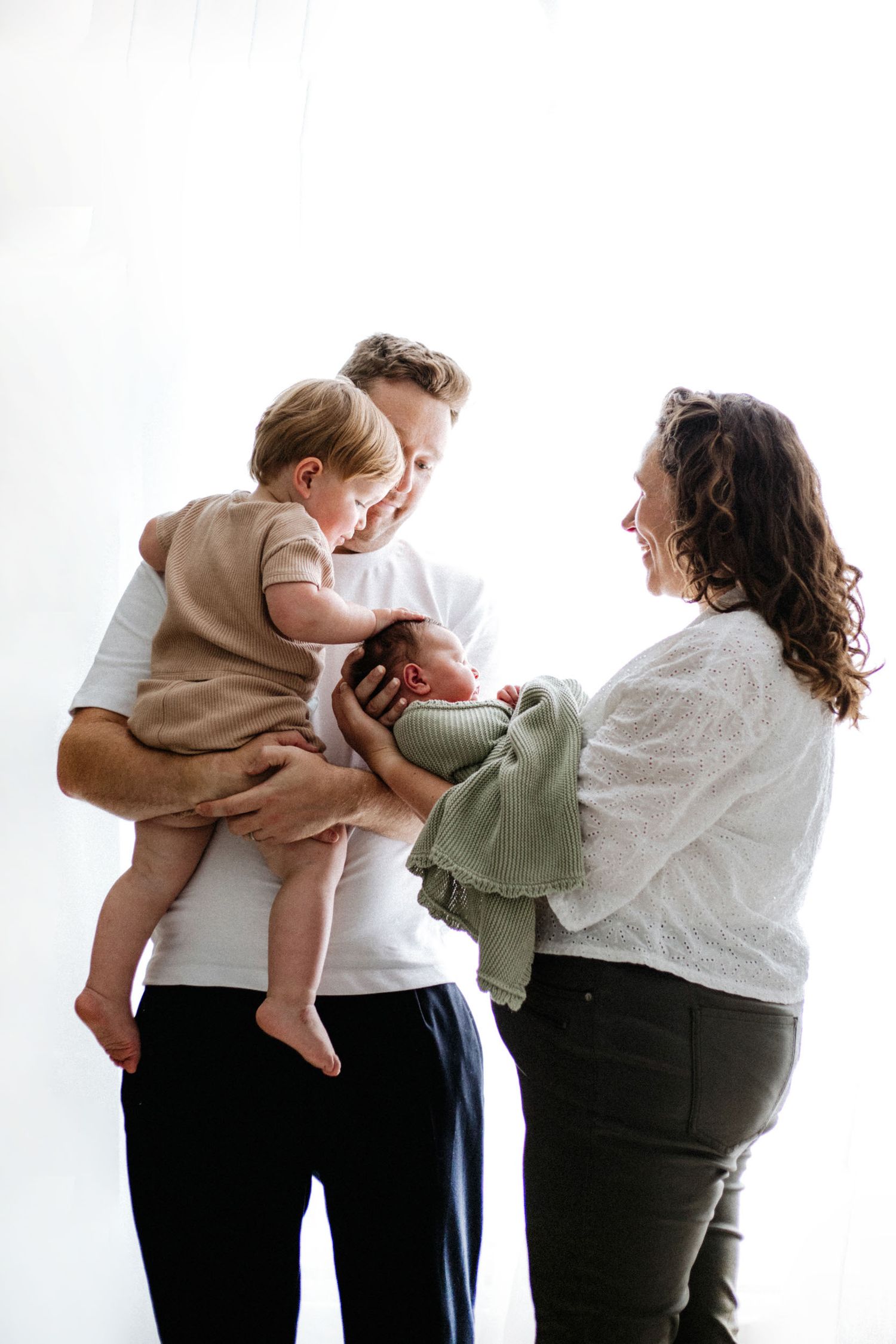 A family of four shares a tender moment, with parents holding a toddler and swaddled newborn baby in a bright studio.