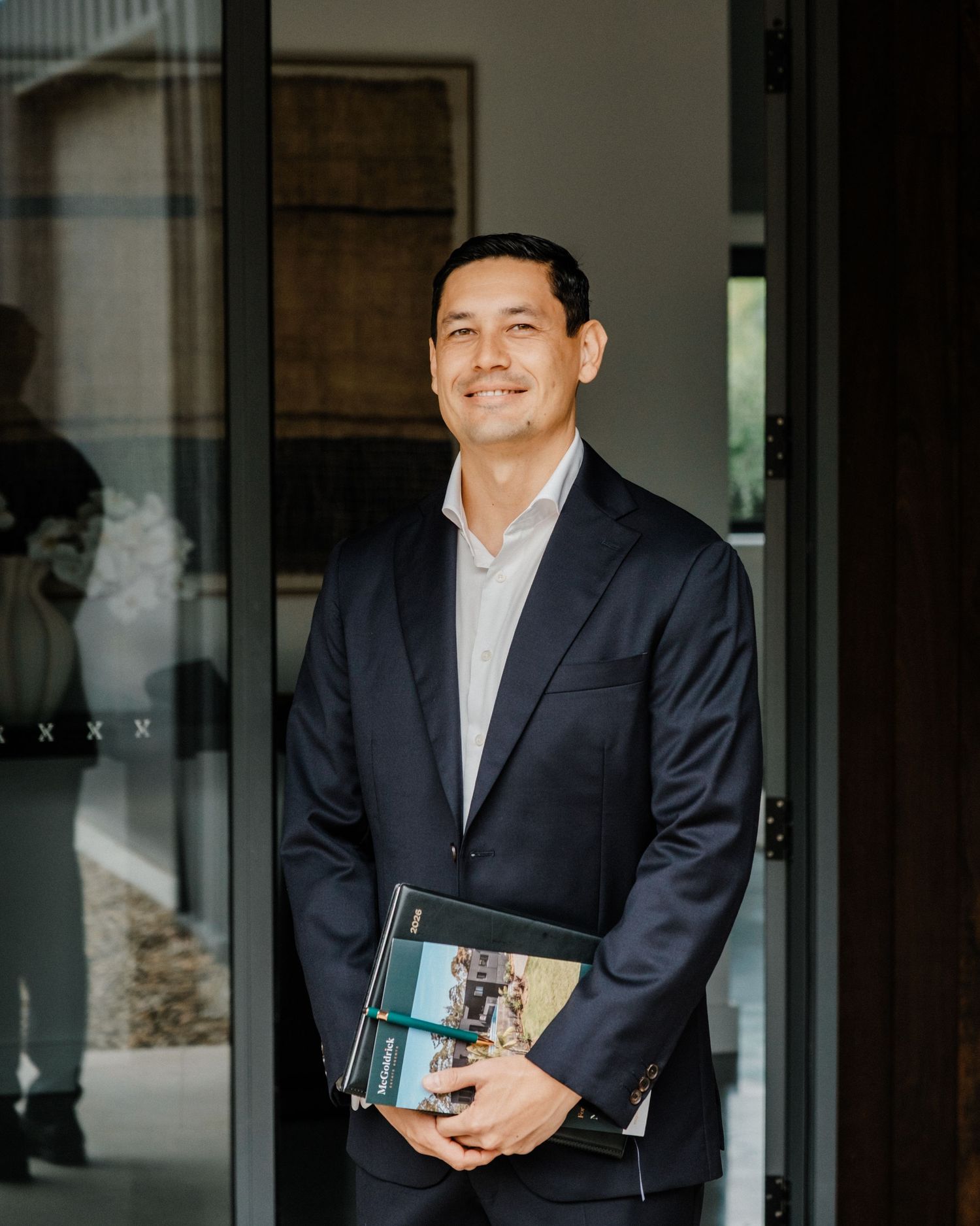 Professional man in navy suit holding documents stands confidently in modern glass doorway.