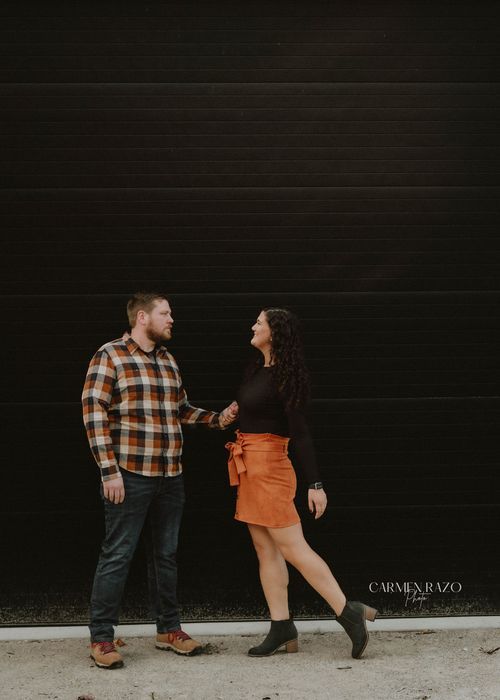 Couple embracing outdoors in autumn park, woman in black top and orange skirt, man in plaid flannel shirt and jeans.