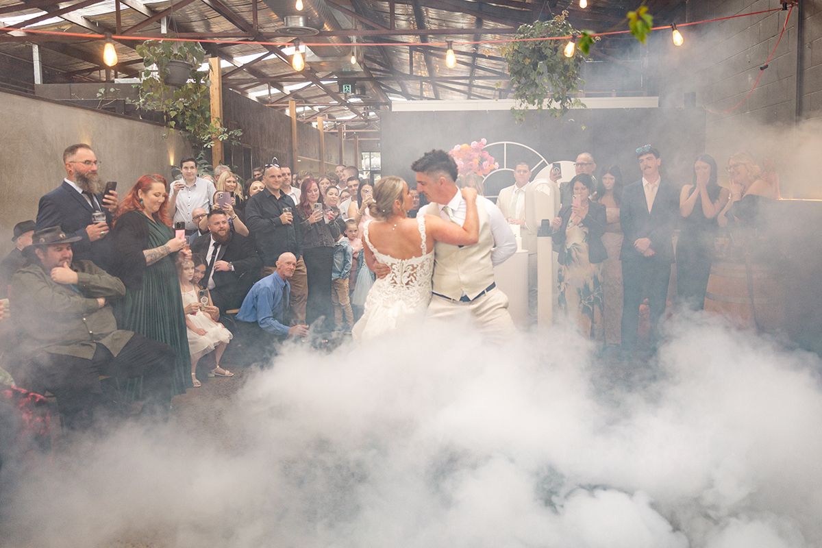 Bride and groom share first dance surrounded by dramatic fog machine smoke at a rustic warehouse wedding reception.