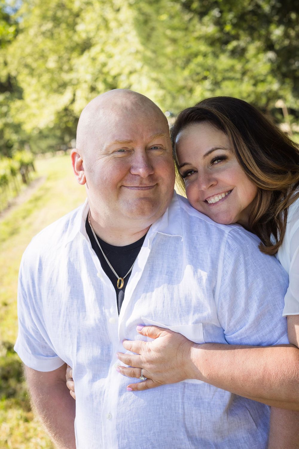 A smiling couple poses outdoors, woman hugging man from behind near lush green trees on a sunny day.