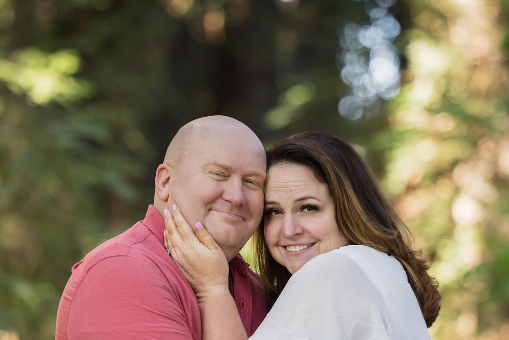 A smiling couple poses outdoors among trees, with a woman in white embracing a bald man in a pink shirt.