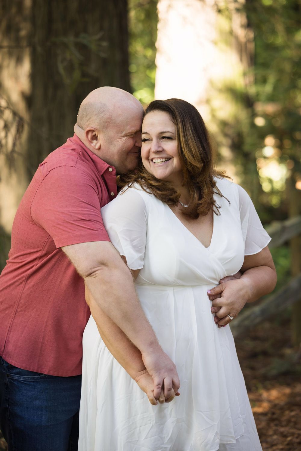 A bald man in a red shirt embraces a smiling woman in a white dress in a sunlit forest setting.
