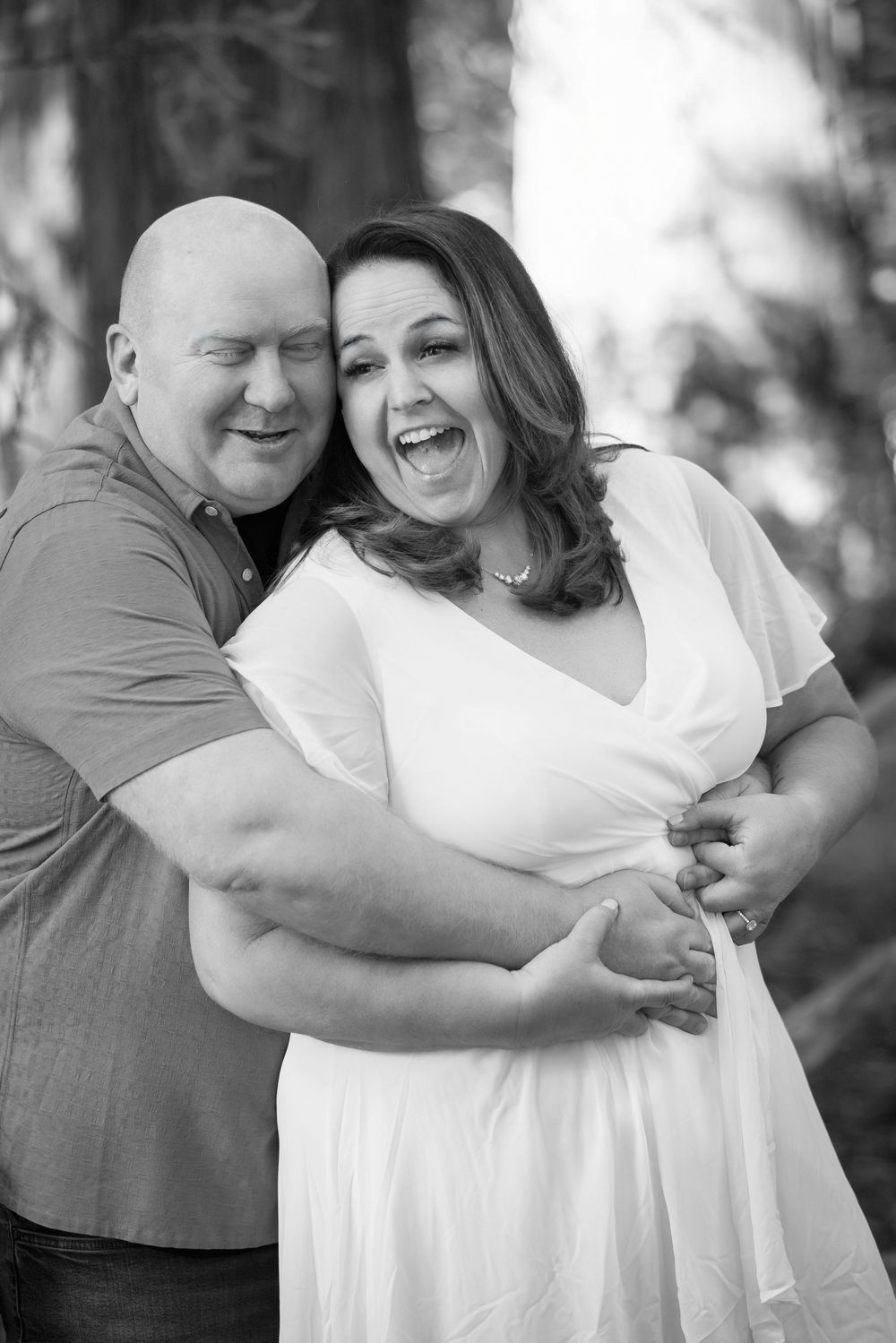 Black and white photo of a happy couple embracing and laughing outdoors among trees.