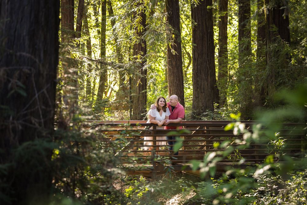 Couple standing on a wooden bridge surrounded by tall redwood trees in a lush forest.