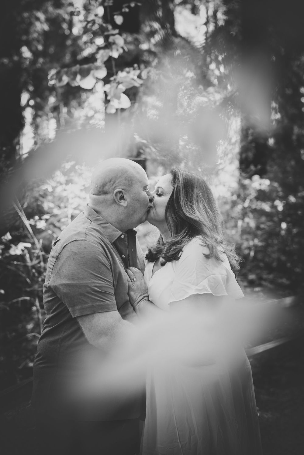 Black and white photo of a couple sharing a tender kiss outdoors surrounded by lush green foliage.