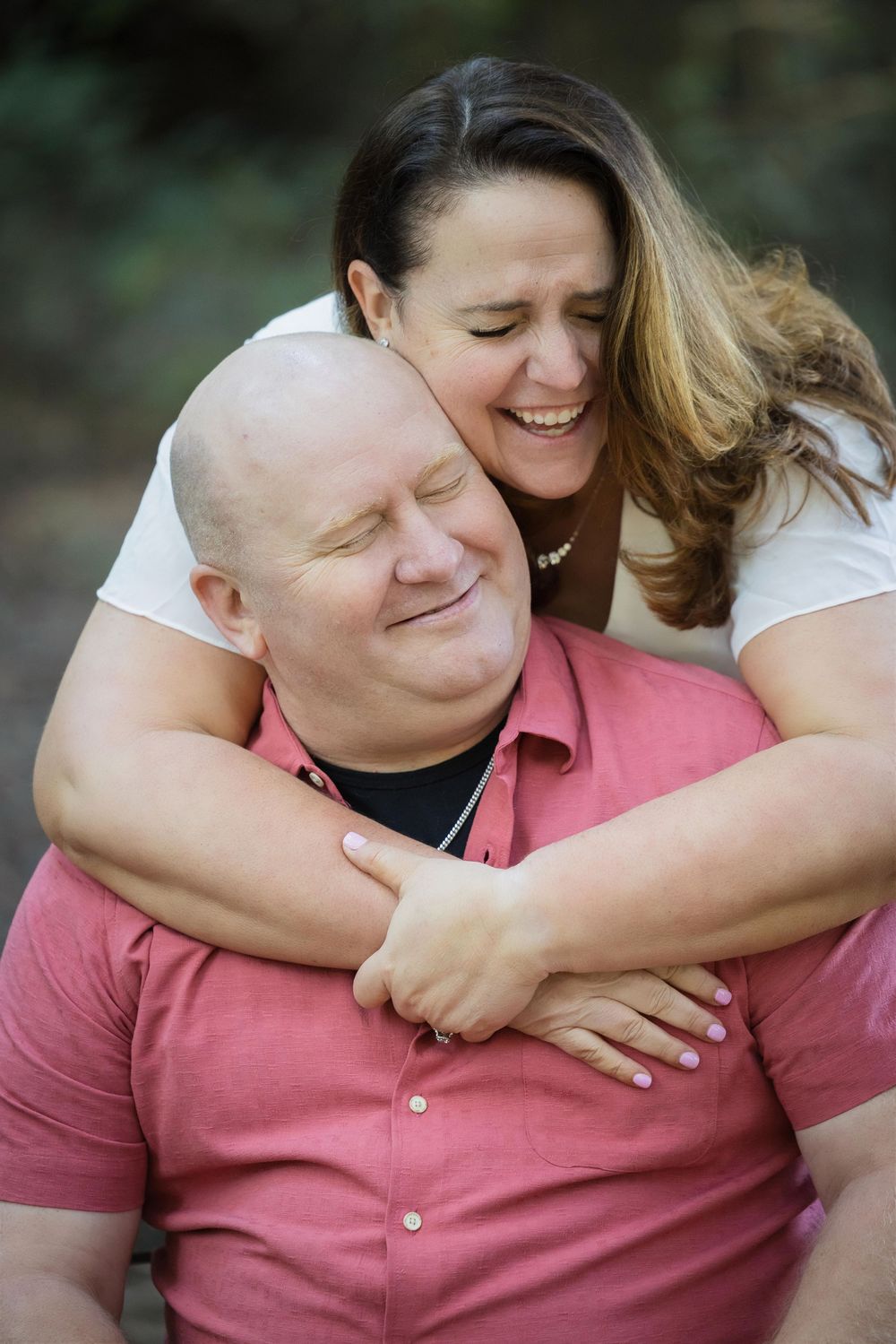 A woman hugs a bald man from behind, both smiling joyfully outdoors in a candid couples portrait.