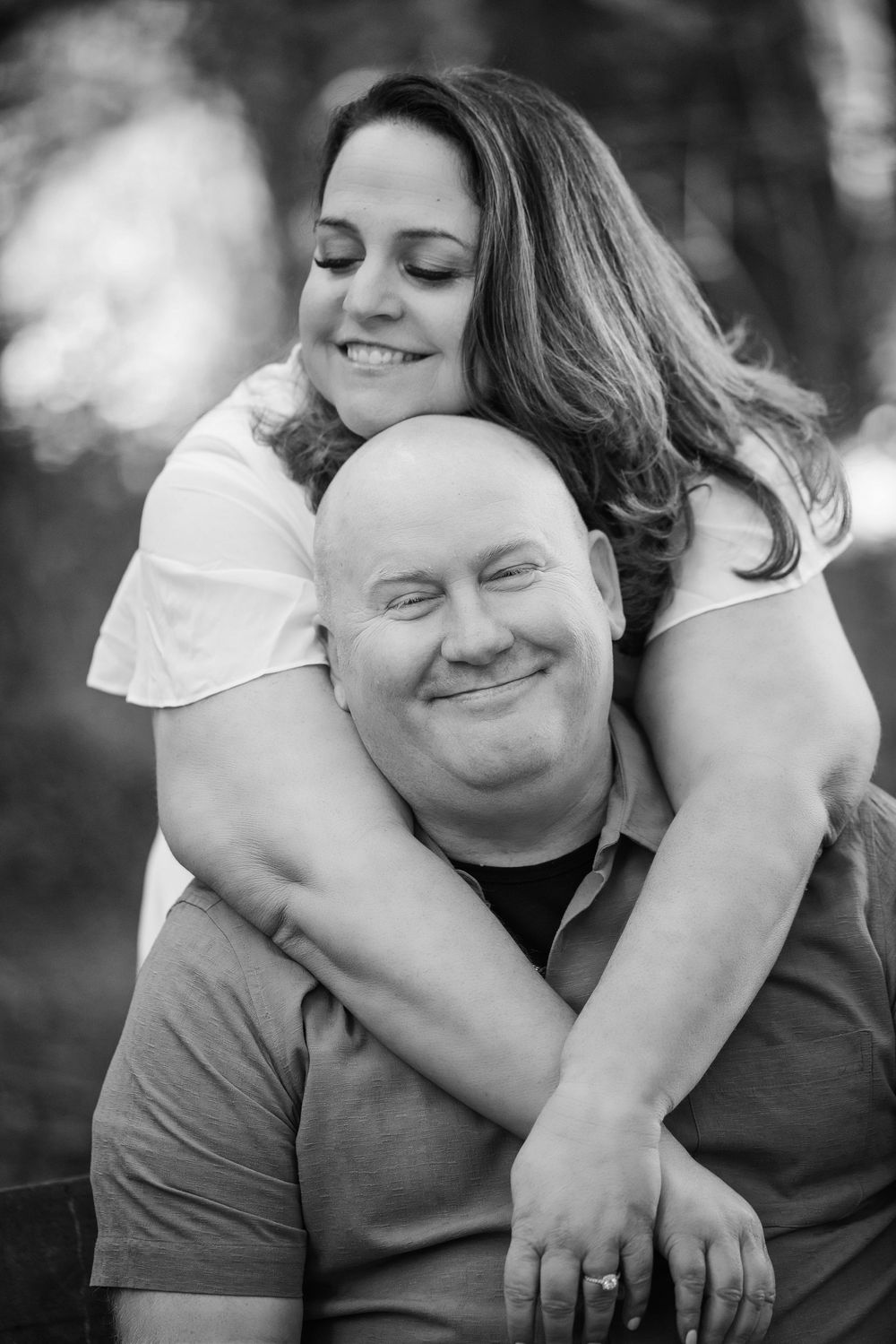 Black and white photo of a smiling woman hugging a bald man from behind outdoors, both appearing joyful.