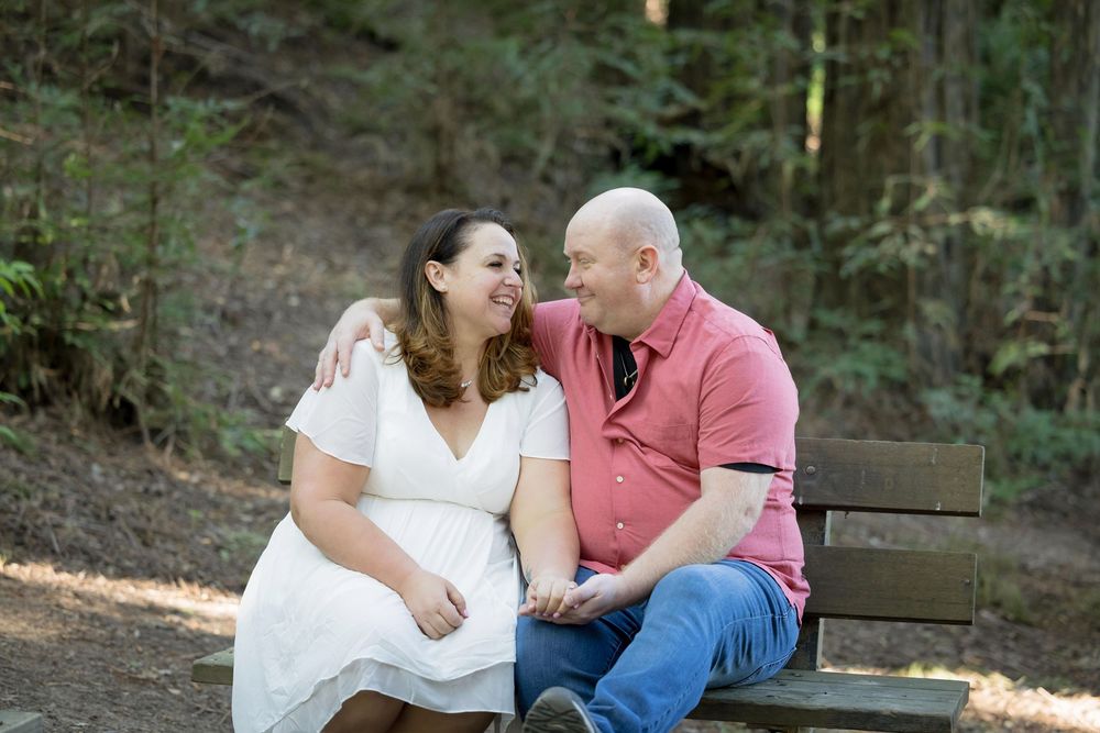 Couple sitting on a park bench smiling at each other, woman in white dress and man in pink shirt, surrounded by trees.