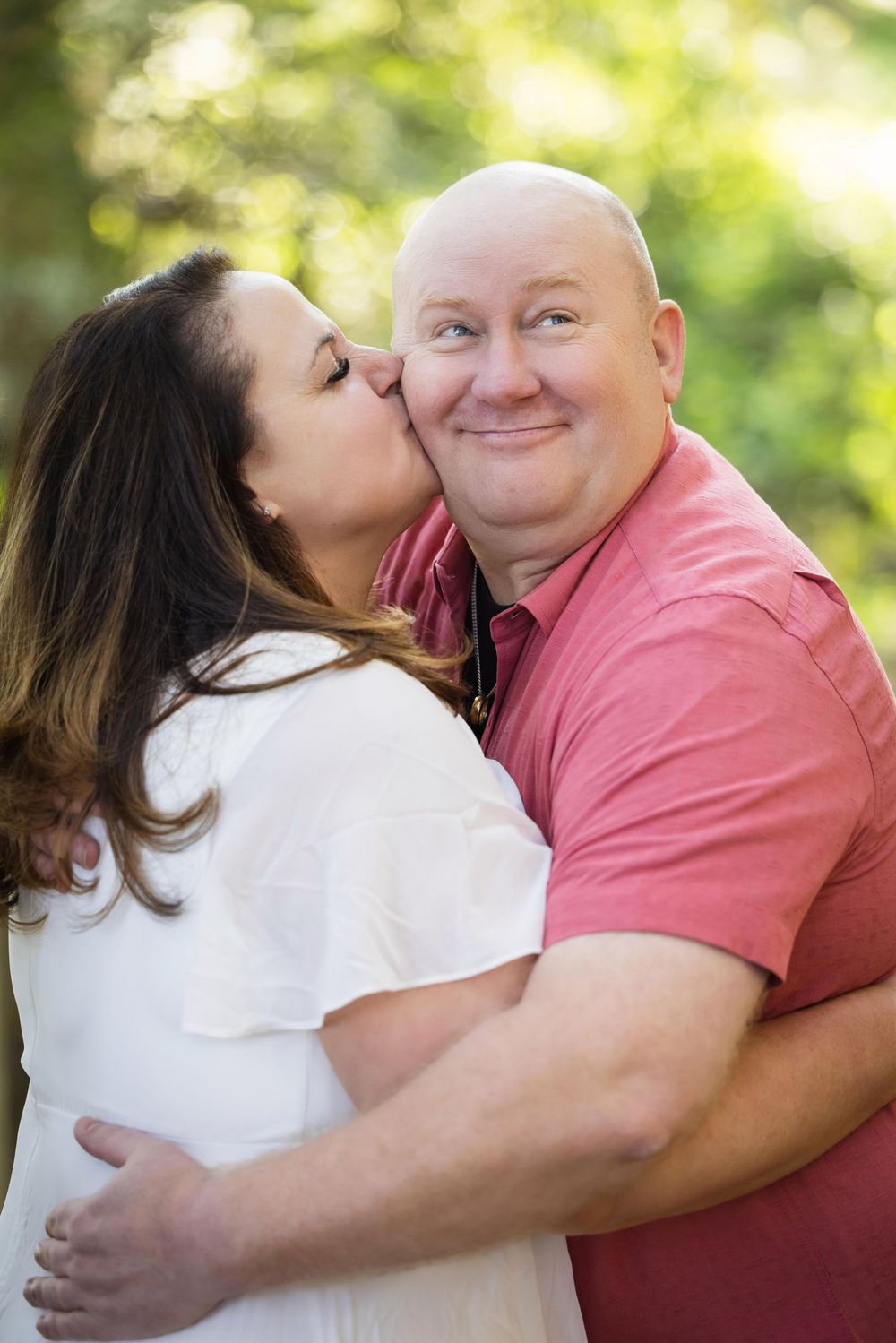 A woman in a white dress kisses a smiling bald man in a pink shirt outdoors with green foliage behind them.