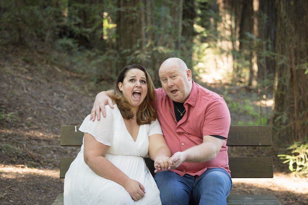 Couple sitting on a bench in a forest, laughing and making surprised expressions, woman in white dress, man in pink shirt.