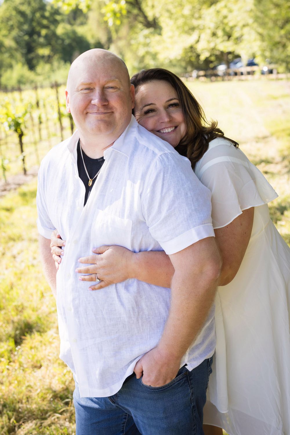 A smiling couple in white shirts poses outdoors in a vineyard on a sunny day.
