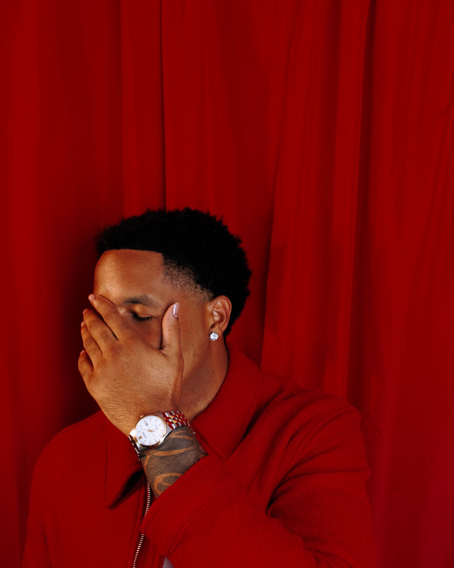 A young man in a red jacket covers his face with his hand, wearing a watch, against a red curtain backdrop.