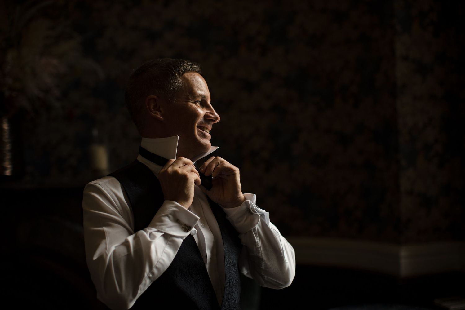A smiling groom in a white shirt and vest adjusts his bow tie while getting ready for his wedding.