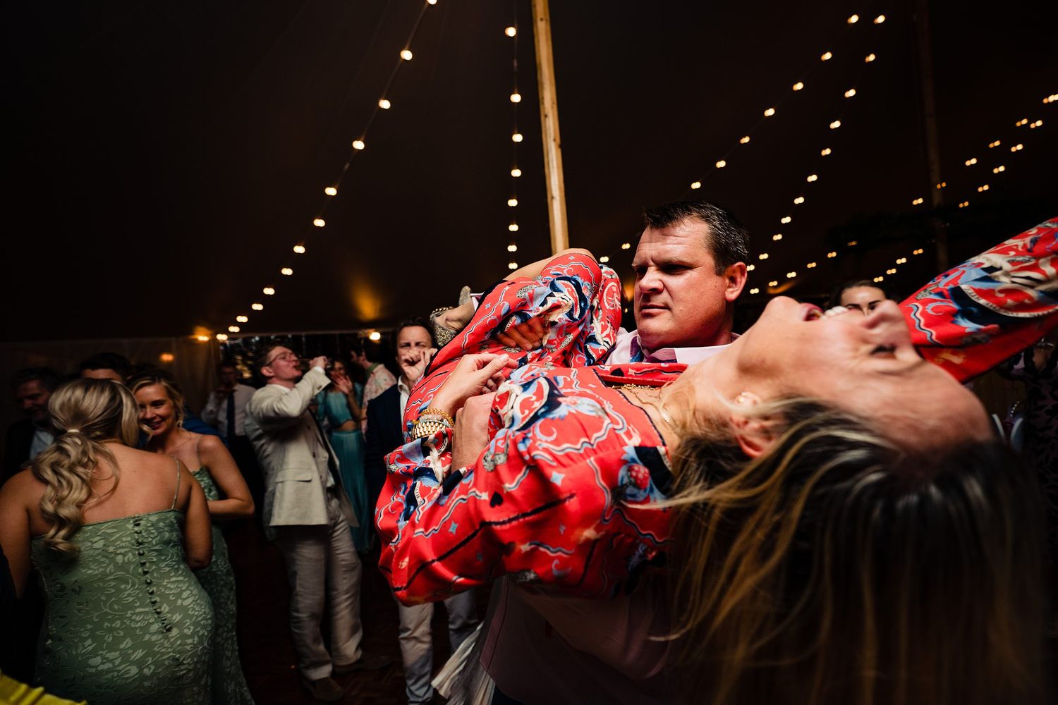 Guests dancing energetically at a wedding reception under warm string lights in a festive tent venue.