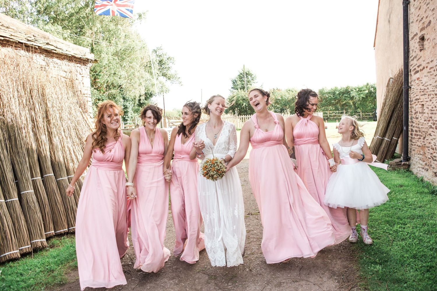 Bride in white gown walks with six bridesmaids in pink dresses and flower girl outside rustic barn venue.