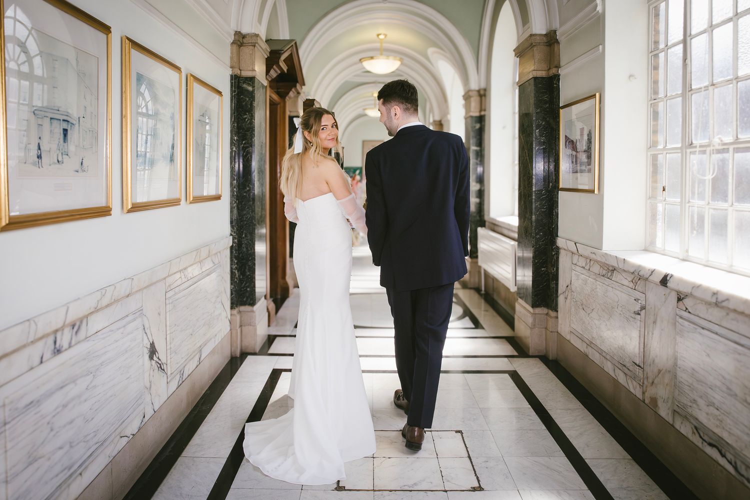 Black and white photo of a bride and groom walking hand in hand down an elegant arched hallway on their wedding day.