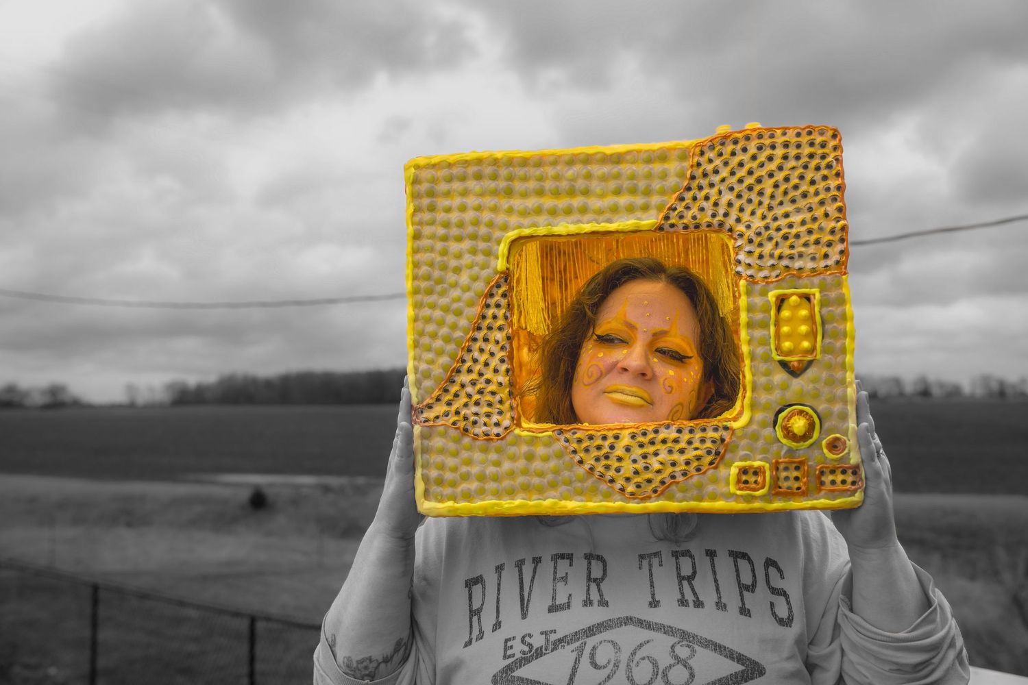 Woman holding a yellow decorative frame in front of her face outdoors, wearing a River Trips 1968 sweatshirt.