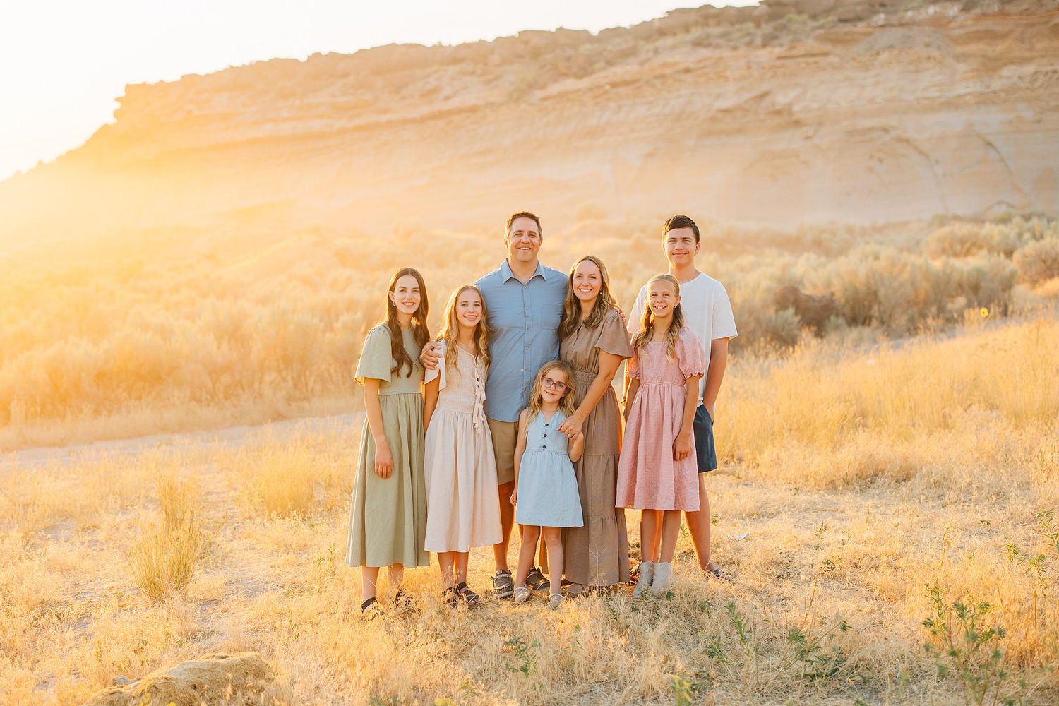 Family of seven posing outdoors at golden hour with a rocky hillside and dry grass in a warm sunset landscape.