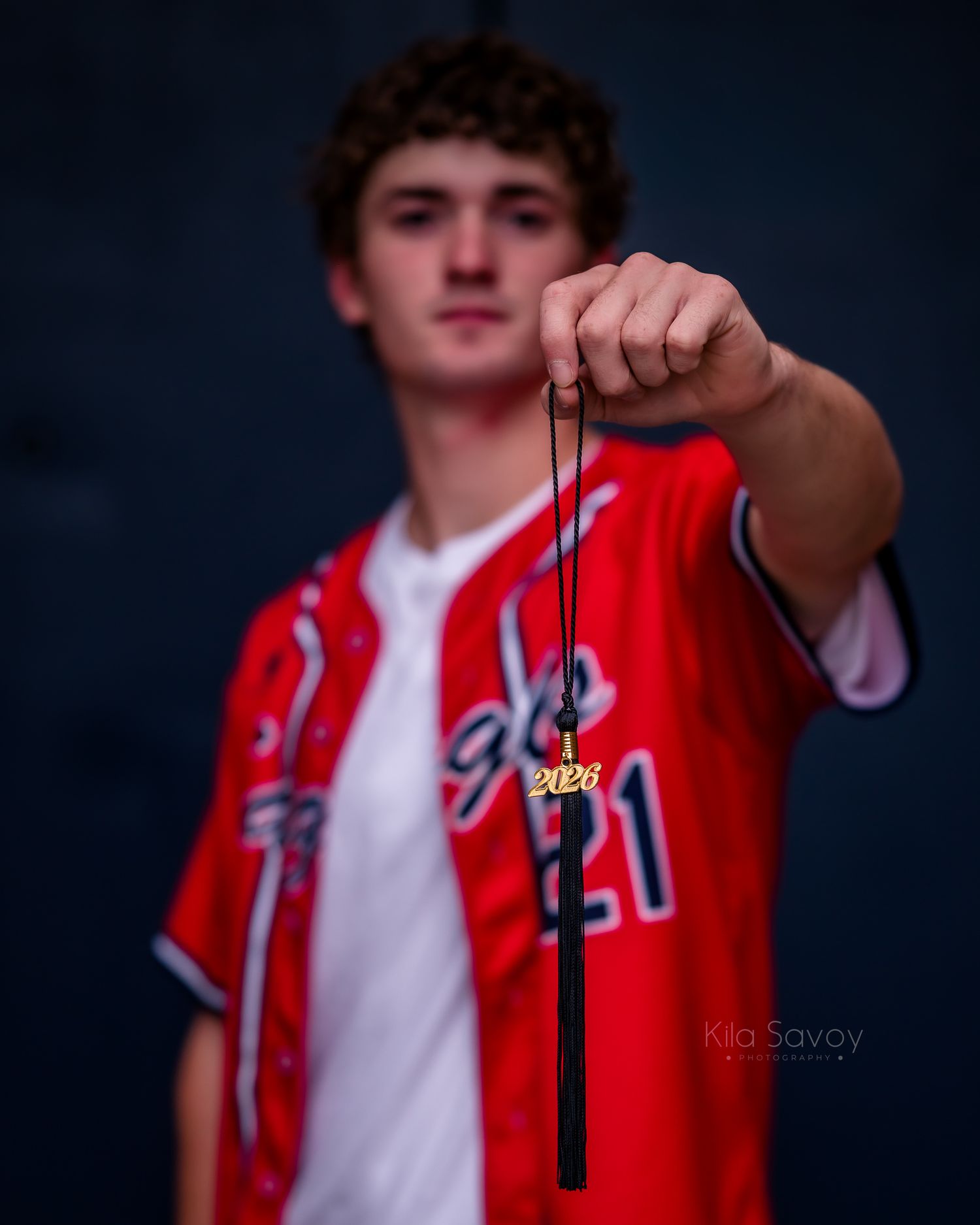 A young man in a red baseball jersey holds up a black graduation tassel with a 2026 charm toward the camera.