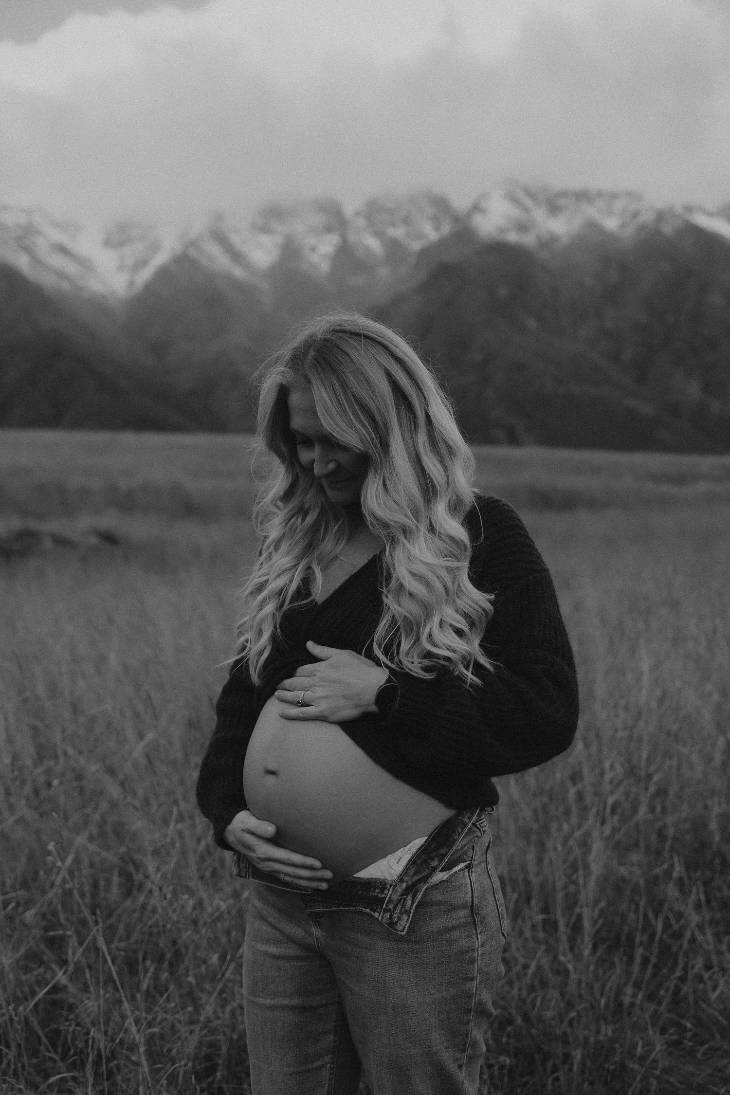 Couple sharing a tender maternity moment in a golden field with snow-capped mountains backdrop.
